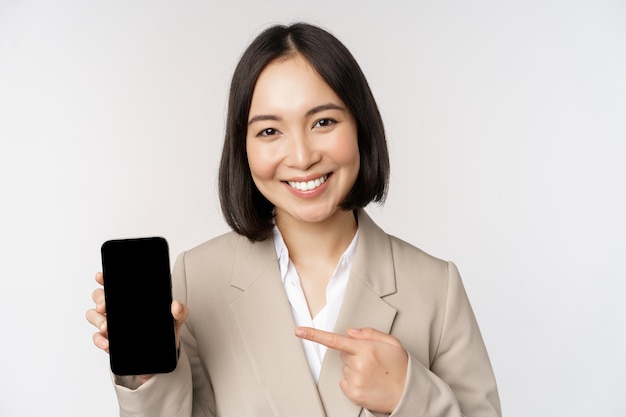 Portrait of corporate asian woman showing smartphone app interface mobile phone screen standing over white background