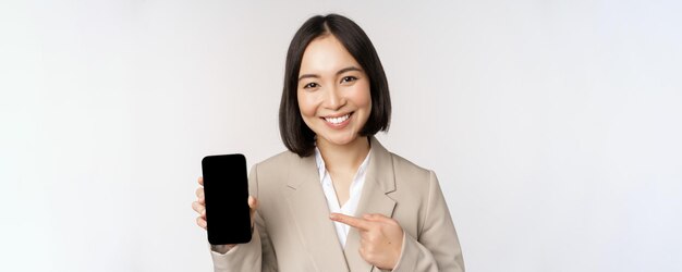 Portrait of corporate asian woman showing smartphone app interface mobile phone screen standing over white background