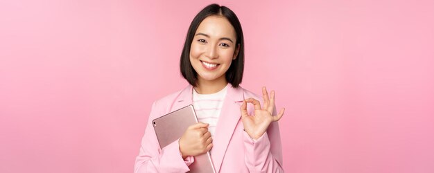 Portrait of corporate woman girl in office in business suit holding digital tablet showing okay recommending company standing over pink background
