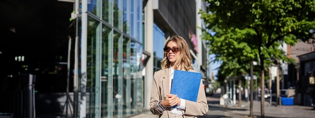 Free photo portrait of corporate woman in sunglasses and beige suit holding blue folder with office documents