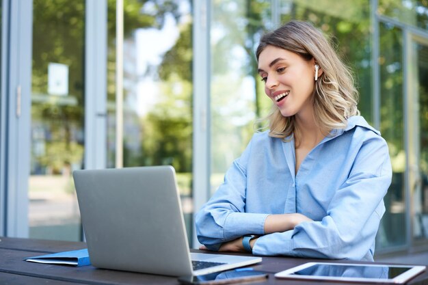 Portrait of corporate woman working outside office on remote sitting with laptop wireless earphones