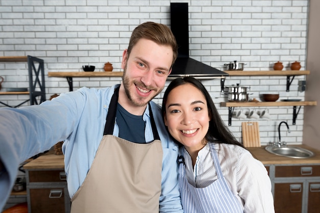 Portrait of couple taking selfie wearing apron in kitchen