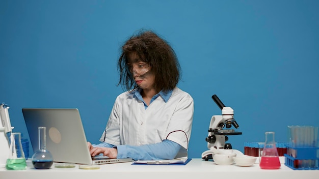 책상에서 노트북 작업을 하는 미친 여성 화학자의 초상화, 스튜디오에서 웃기고 멍청한 모습. 미친 미친 과학자는 컴퓨터를 사용하고 어리석은 어리석은 표정을 짓고 머리를 헝클어뜨립니다.