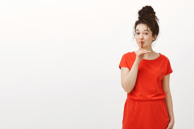 Portrait of creative cheerful Caucasian woman with curly hair combed in bun, smiling curiously while saying shh