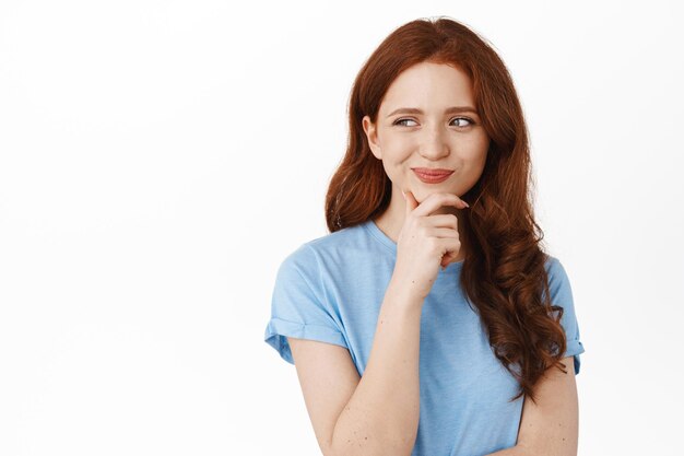 Portrait of creative redhead girl student thinking, wearing summer clothes, looking aside at advertisement thoughtful, touch chin and pondering, smiling pleased with good idea, white background.