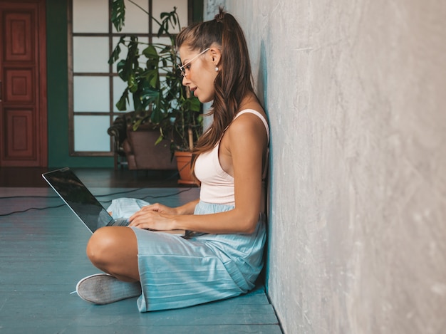 Portrait of creative young smiling woman in sunglasses. Beautiful girl sitting on the floor near gray wall in the interior. Model using notebook. Female dressed in hipster clothes