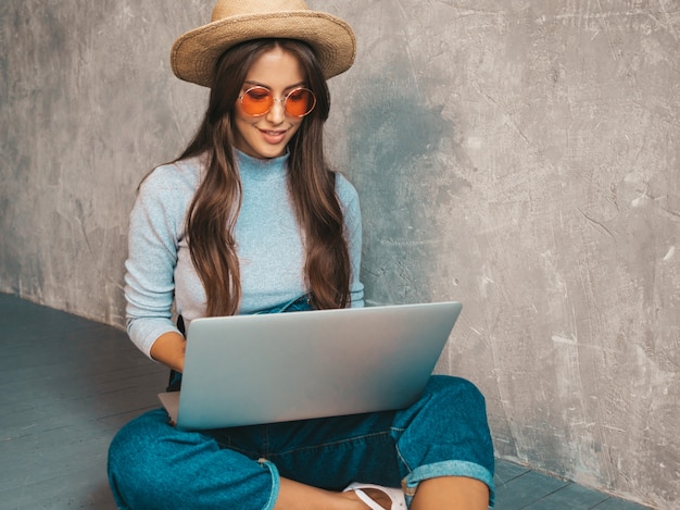 Portrait of creative young smiling woman in sunglasses. Beautiful girl sitting on floor near gray wall..Typing and searching information