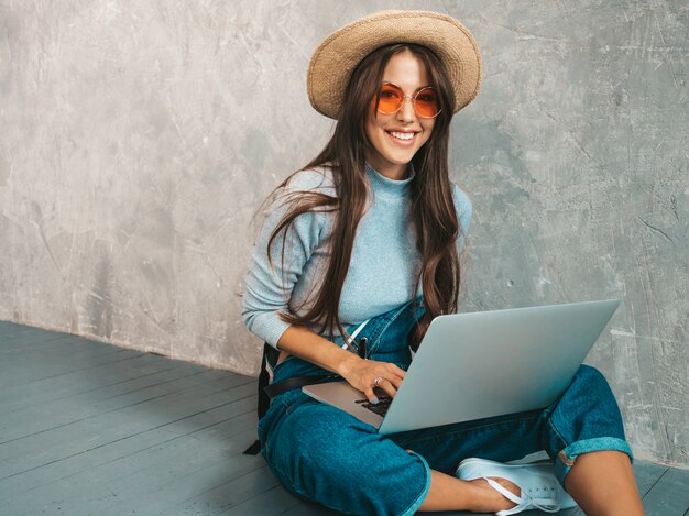 Portrait of creative young smiling woman in sunglasses. Beautiful girl sitting on the floor near gray wall. 