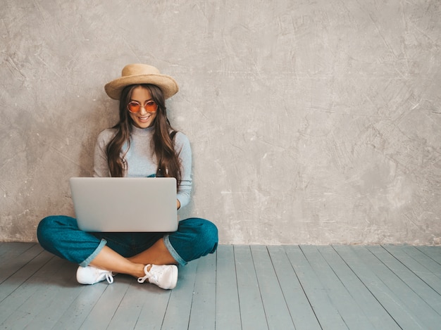 Portrait of creative young smiling woman in sunglasses. Beautiful girl sitting on the floor near gray wall. 