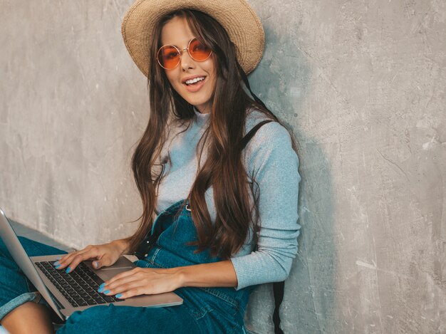 Portrait of creative young smiling woman in sunglasses. Beautiful girl sitting on the floor near gray wall. 