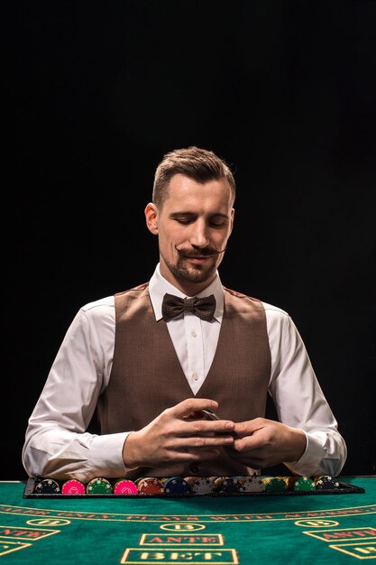 Portrait of a croupier is holding playing cards, gambling chips on table. Black background. A young male croupier in a shirt, waistcoat and bow tie is waiting for you at the blackjack table