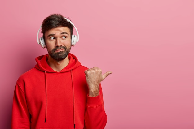 Foto gratuita il ritratto dell'uomo barbuto curioso punta il pollice nello spazio vuoto a destra, indossa l'auricolare stereo e una felpa casual rossa, dimostra qualcosa di interessante, isolato sul muro rosa pastello.