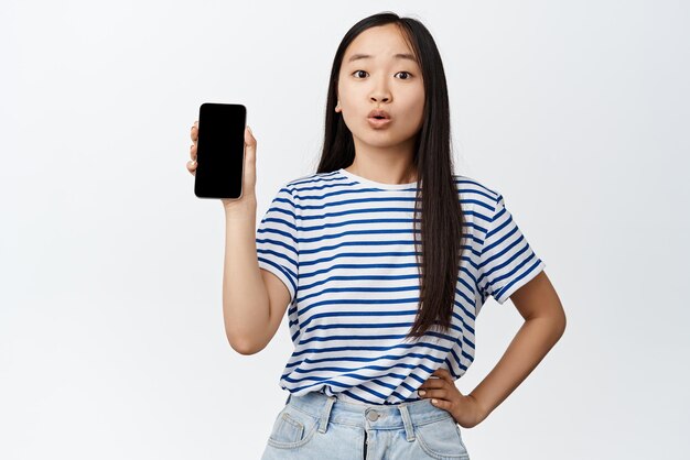 好奇心旺盛な女の子の肖像画は、面白いアプリが携帯電話を保持し、スマートフォンの画面の白い背景に何かをお勧めしていることを示しています