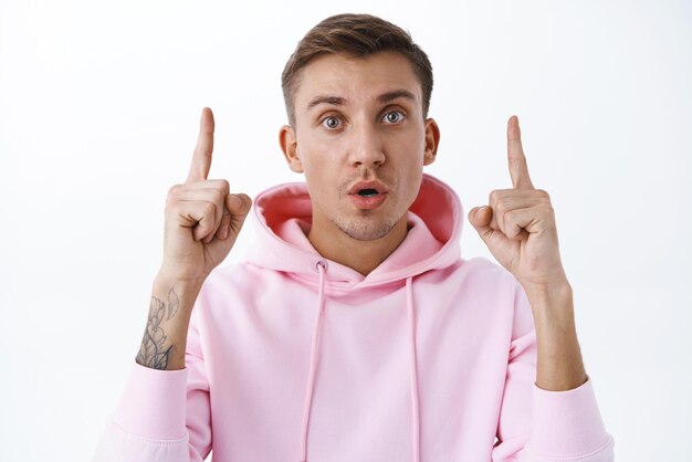 Portrait of curious handsome blond guy in pink hoodie pointing fingers up stare camera open mouth amused found out great promo asking question being interested and intrigued white background