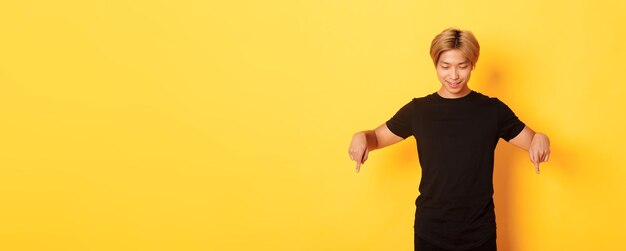 Free photo portrait of curious happy blond asian guy looking and pointing fingers down with pleased smile yello