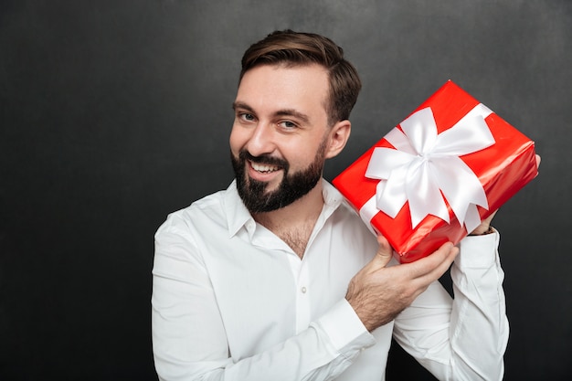 Portrait of curious man shaking red box gift wrapped, and trying to recognize what's inside over dark gray wall