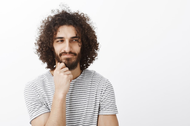 Portrait of curious playful handsome male with curly hair, looking aside and smirking, holding hand on beard while thinking and having great plan