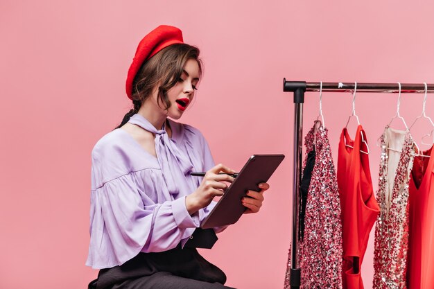 Portrait of curly girl with red lipstick taking notes in tablet on pink background with dressees.