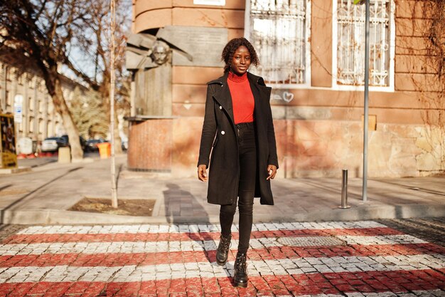 横断歩道を歩いているファッショナブルな黒いコートと赤いタートルネックを身に着けている縮れ毛のアフリカの女性の肖像画