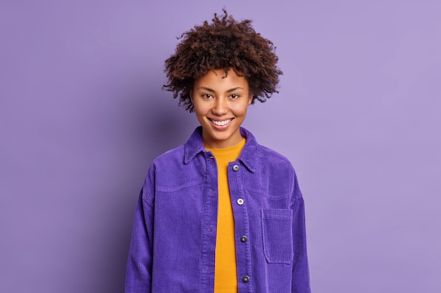 Portrait of curly haired beautiful woman smiles happily wears fashionable purple jacket in one tone with background feels delighted poses glad indoor talks to coworker. Positive emotions concept