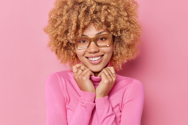 Free photo portrait of curly haired woman smiles gently keeps hands on collar of jumper expresses positive emotions hears good news wears spectacles isolated over pink background. happy emotions concept