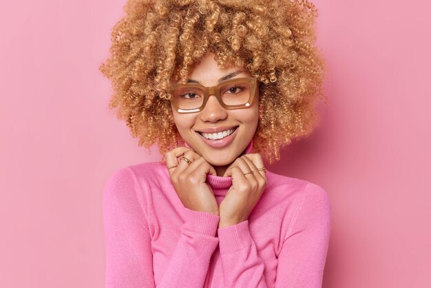 Portrait of curly haired woman smiles gently keeps hands on collar of jumper expresses positive emotions hears good news wears spectacles isolated over pink background. Happy emotions concept
