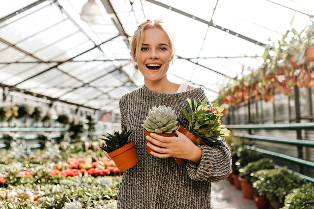 鉢植えの植物をたくさん持っている灰色のセーターの巻き毛の女性の肖像画。植物店で笑顔のポーズで緑色の目のブロンド。