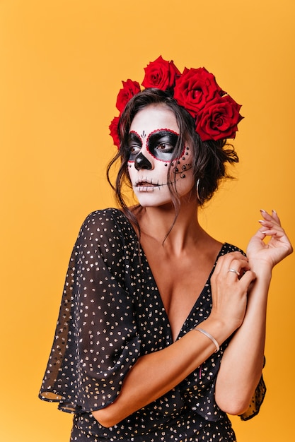 Free photo portrait of curly woman with red big roses in her hair mysteriously looking into distance against wall of orange wall. girl model from mexico with makeup for halloween posing
