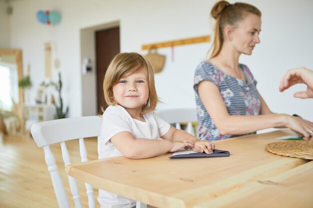 Portrait of cute adorable baby girl in white t-shirt sitting at wooden dining table wiht her mother, learning how to make origami paper plane,  with happy smile. Selective focus