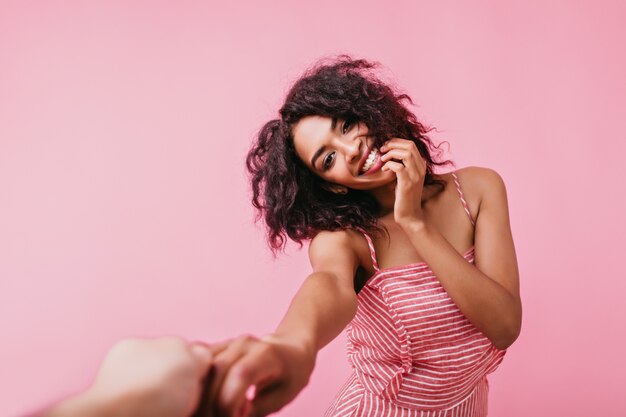 Portrait of cute American girl, sincerely smiling and touching her face. Lady with dark curls holds friend's hand.