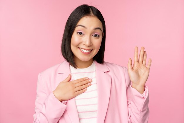 Портрет милой азиатской деловой женщины, поднимающей руку, представляющейся в офисе, кокетливо улыбающейся, стоящей на розовом фоне в костюме