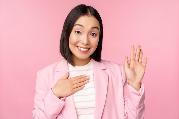 Portrait of cute asian businesswoman raising hand introduce herself in office smiling coquettish standing over pink background in suit