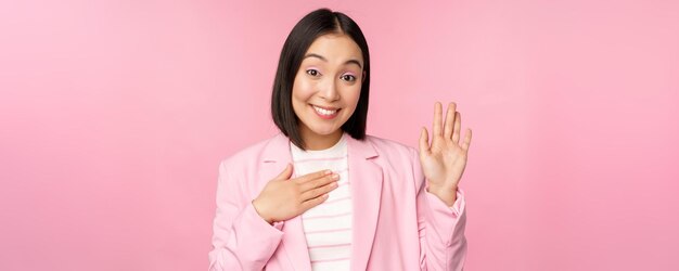 Portrait of cute asian businesswoman raising hand introduce herself in office smiling coquettish standing over pink background in suit