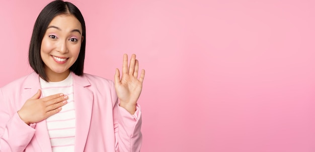 Portrait of cute asian businesswoman raising hand introduce herself in office smiling coquettish standing over pink background in suit