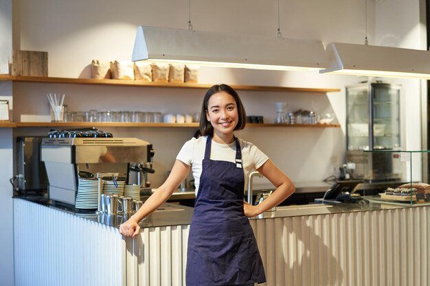 ap를 입고 커피 기계와 카운터 근처에 서있는 귀여운 아시아 여성 바리 스타 카페 직원의 초상화
