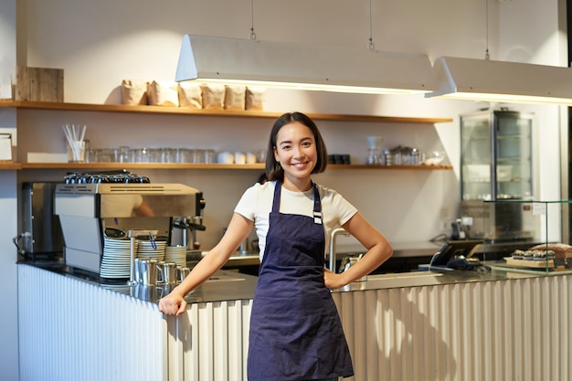 Portrait of cute asian woman barista cafe staff standing near counter with coffee machine wearing ap