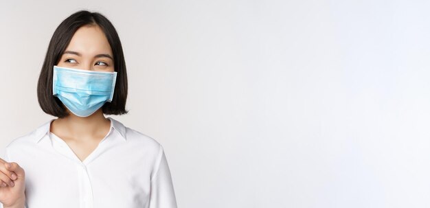 Portrait of cute asian woman in medical face mask coronavirus protection pointing finger left and looking intrigued at empty copy space white background