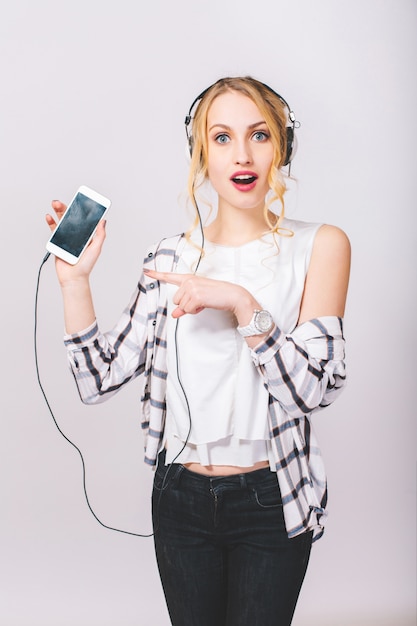 Portrait of cute beautiful blonde girl with surprised face posing, looking. Happy pleased woman showing her smartphone. Wearing stylish outfit.