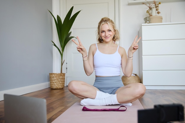 Portrait of cute blond woman shows peace sign smiles records yoga vlog on digital camera workout at