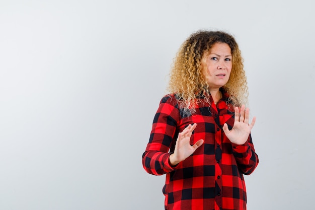 Portrait of cute blonde woman showing stop gesture in checked shirt and looking scared front view