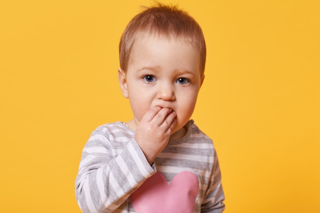 Portrait of a cute brooding girl with short fair hair keeping fingers in her mouth. serious kid stands in front of camera looking straight