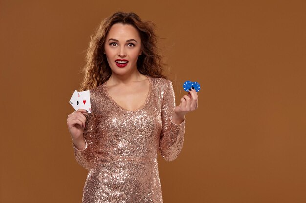 Портрет милой шатенки кавказской девушки на коричневом фоне в золотом коктейльном платье в казино держит в руках пару тузов и фишек. Игорная индустрия, покер, казино