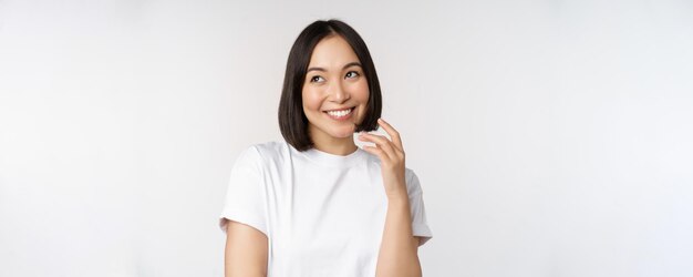スタジオの背景の上に白いTシャツに立っている思慮深い思考やイメージングsmthを脇に見て笑って笑っているかわいいコケティッシュな女性の肖像画
