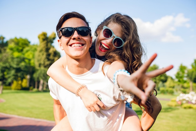 Portrait of cute couple having fun in park. Pretty girl with long curly hair is riding on back of handsome guy. They wear sunglasses and smile to camera.