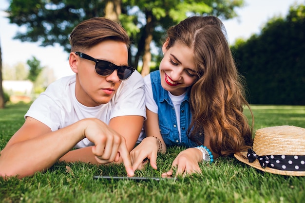 Portrait of cute couple lying on grass in summer park. Girl with long curly hair, red lips is smiling to tablet on grass. Handsome guy in white T-shirt shows on screen.