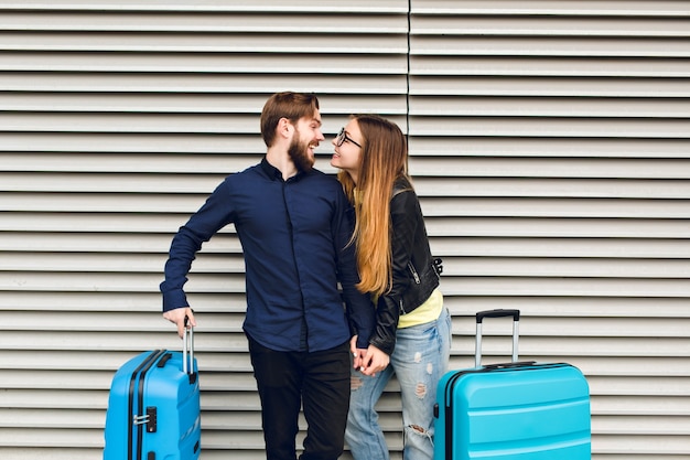 Portrait of cute couple  standing on gray  striped background.  She has long hair, glasses,  yellow sweater, jacket. He wears black shirt, beard. They are  hugging and smilinhg to each other.