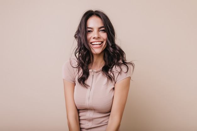 Portrait of cute dark-haired girl with snow-white smile. Brown-eyed lady in beige top looking