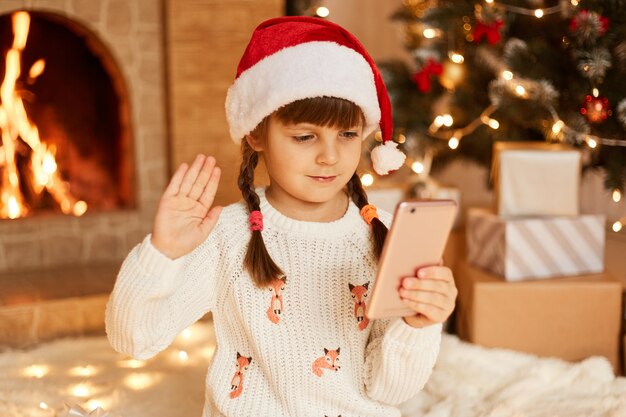 Portrait of cute female child wearing white sweater and santa claus hat, having video call, waving hand to cellphone camera, posing in festive room with fireplace and Xmas tree.