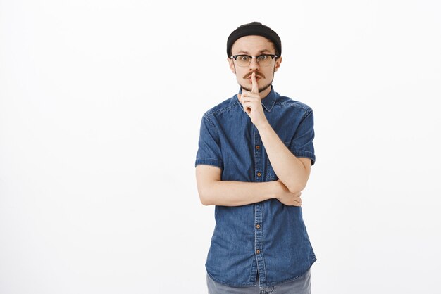 Portrait of cute friendly-looking nervous european man in glasses and black beanie making shush gesture with index finger over mouth and raised eyebrows asking keep secret