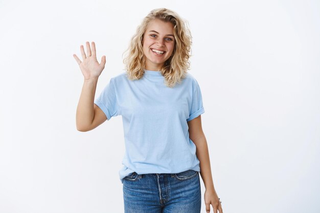 Portrait of cute friendly and positive nice girl with blond hair and blue eyes smiling pleasant as waving palm in greeting or hello gesture being welcoming and nice over white wall